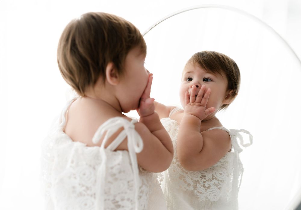Baby in white dress looking at reflection in mirror during photo session.