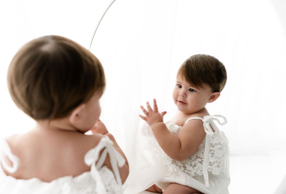 Baby in white lace dress looking at reflection in studio mirror setup.