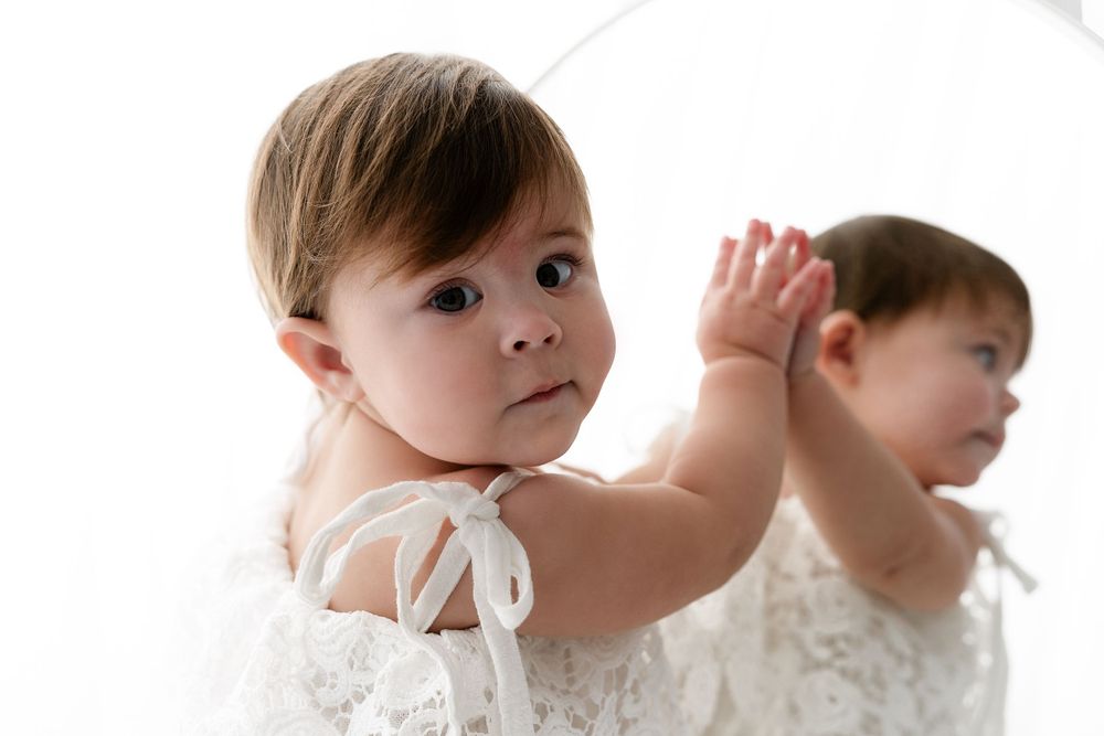 Baby in white dress posing with mirror reflection in minimalist studio setting.