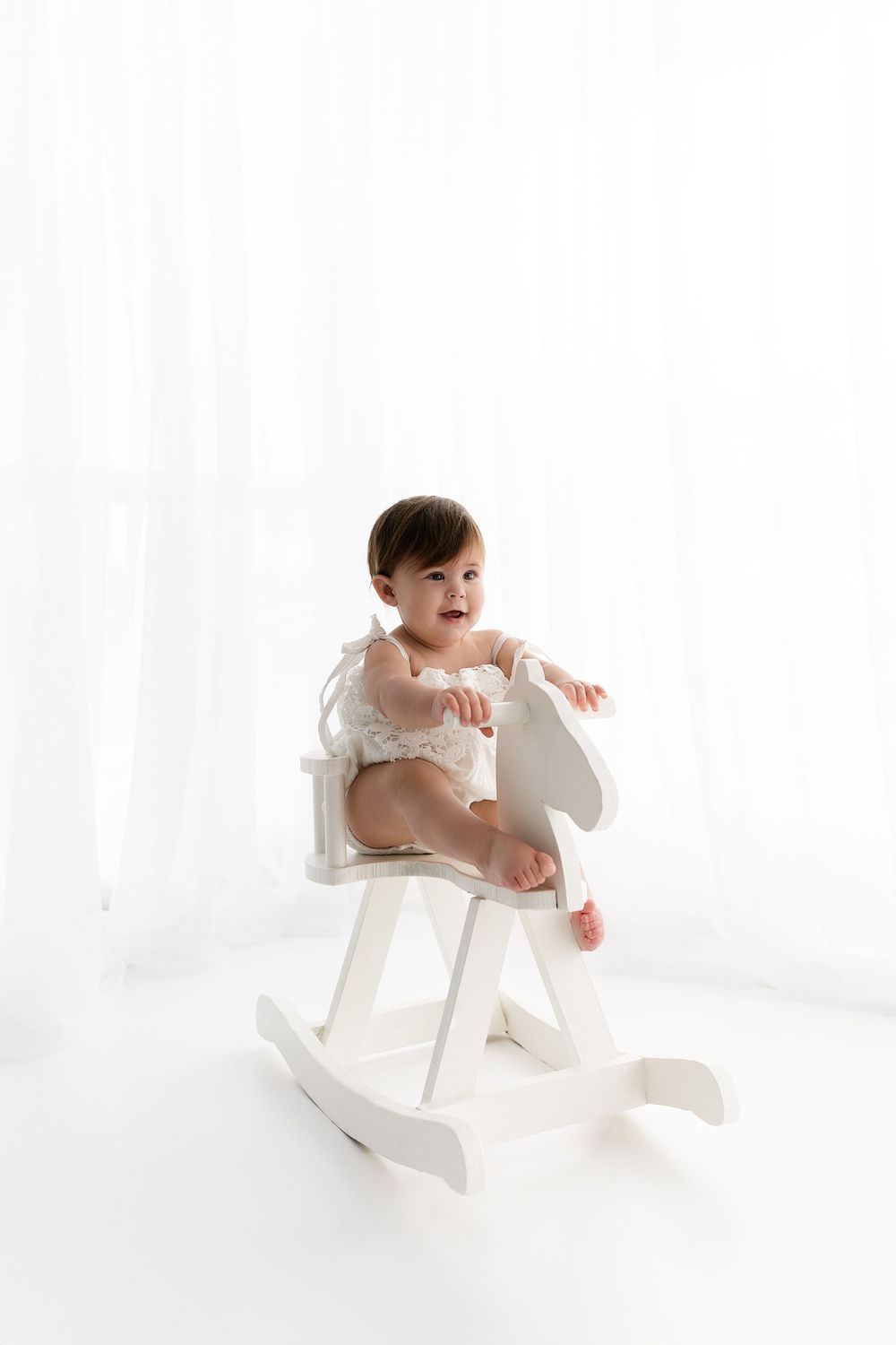Baby in white lace outfit sits on small white rocking horse.