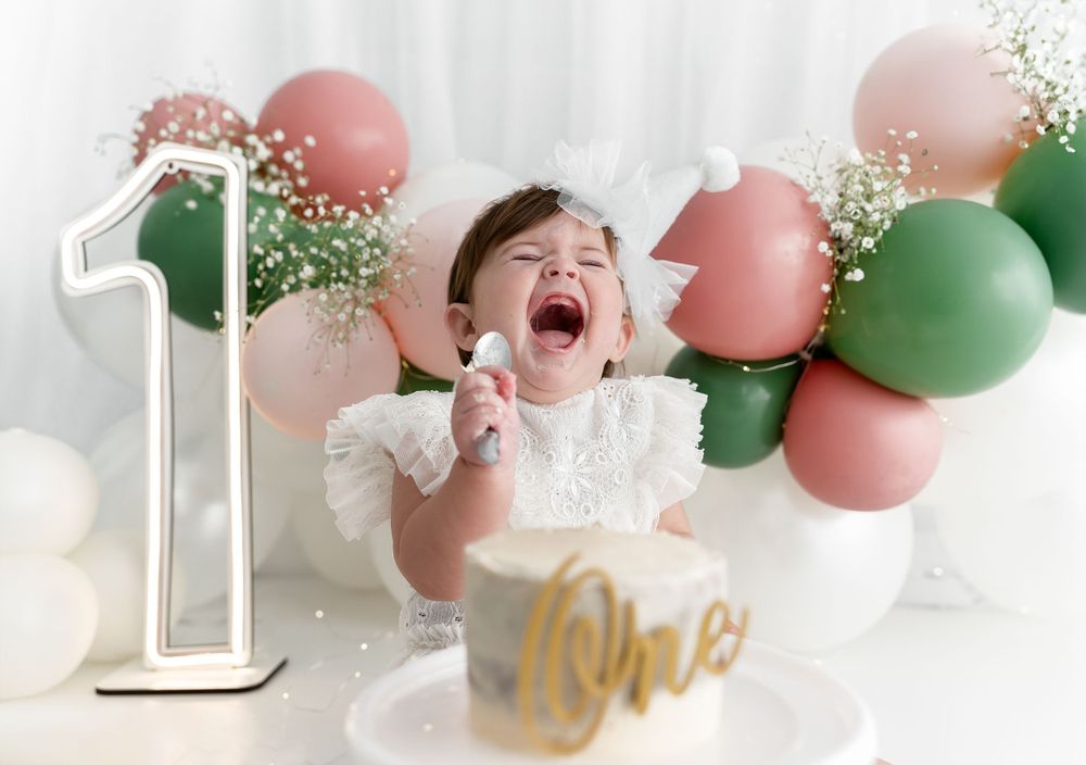 First birthday celebration with balloons, number one sign, and excited baby in white dress.