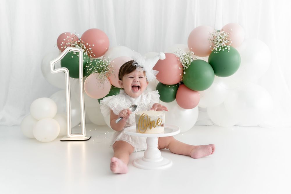 First birthday celebration setup with pink and green balloons, white cake stand, and gold number one decoration.