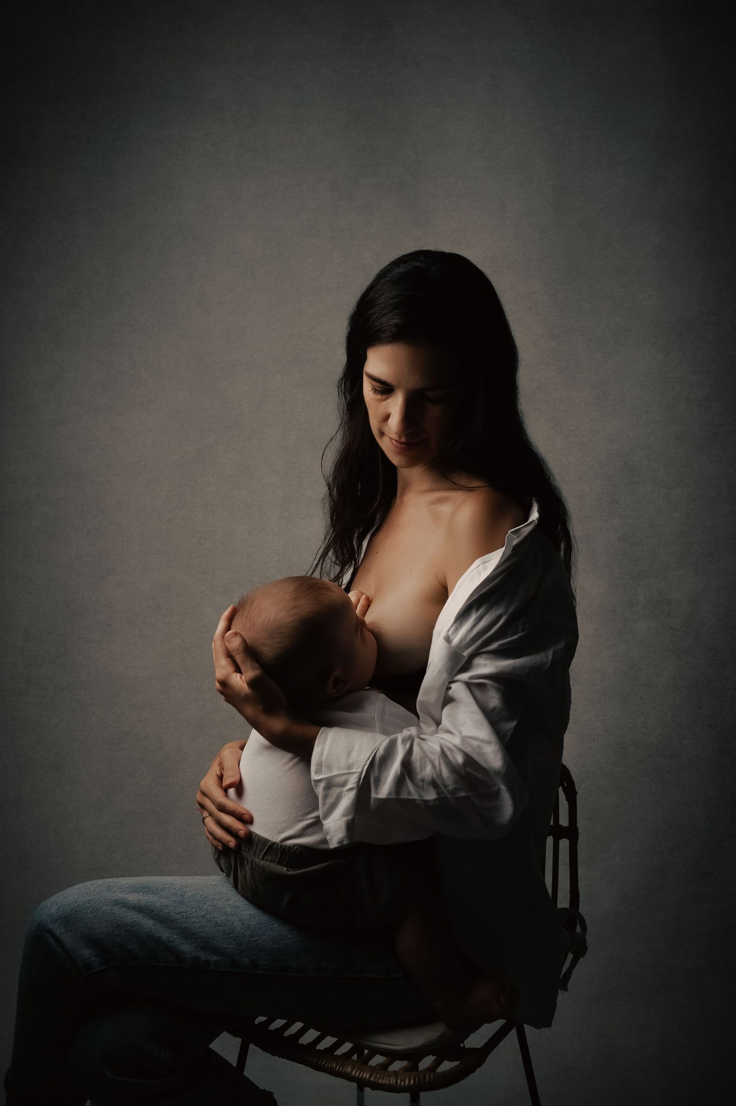 Moody studio portrait of nursing mother in white shirt and jeans against dark background.