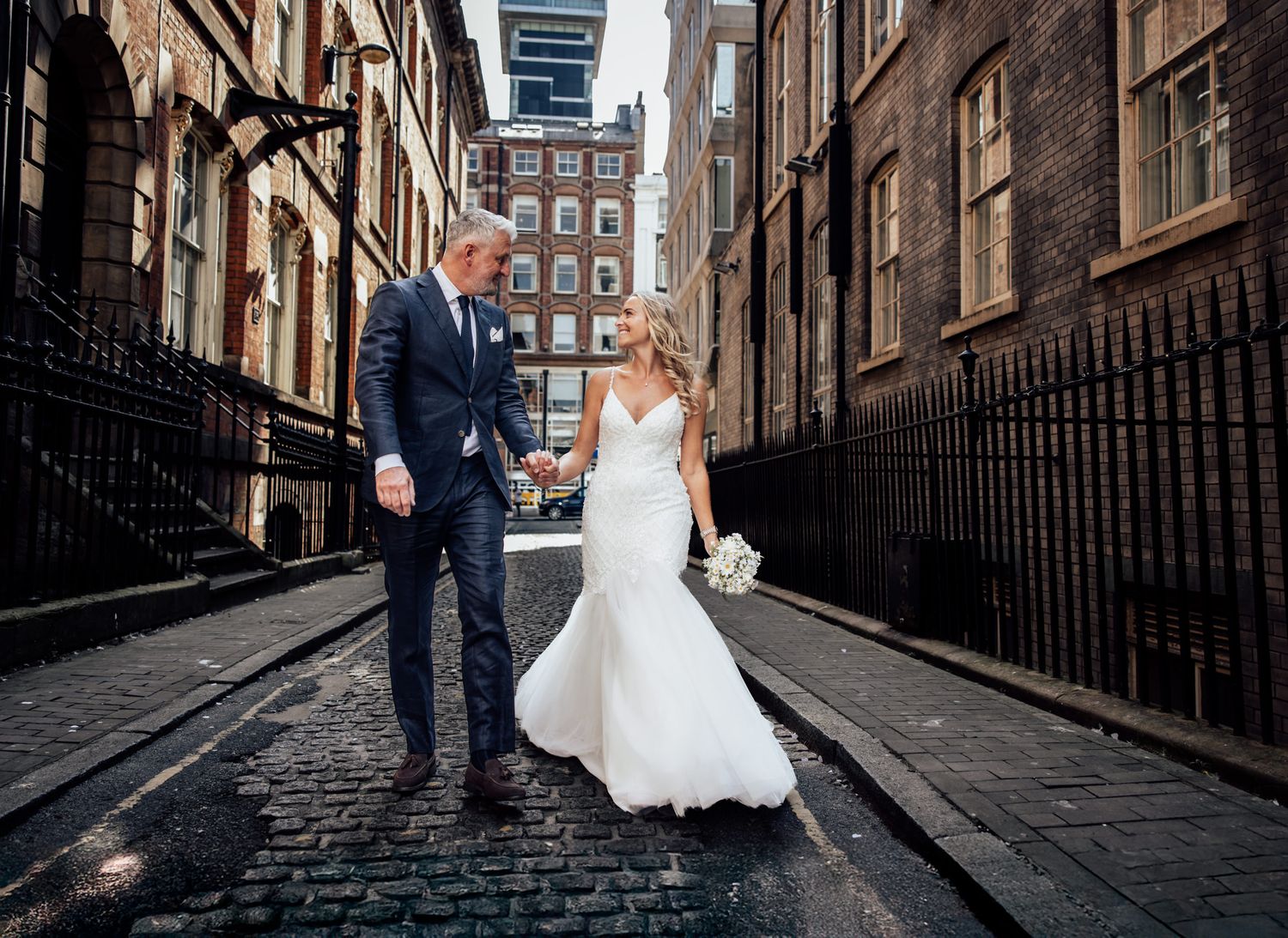 Groom wearing a smart suit holds his brides hand stood in the driveway at Bartle Hall wedding venue in Preston