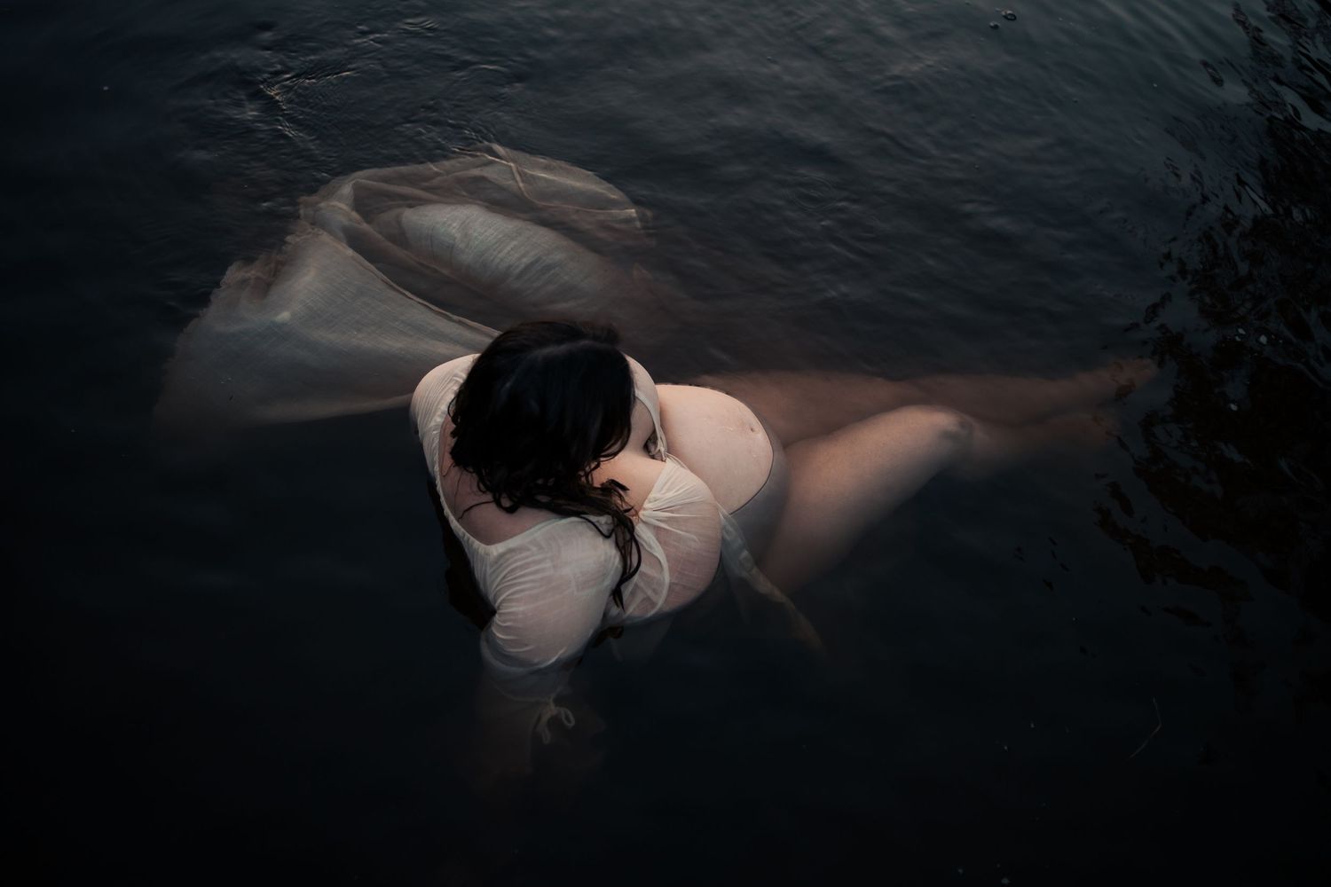 A figure in white floats ethereally in dark water creating a dramatic artistic underwater portrait.