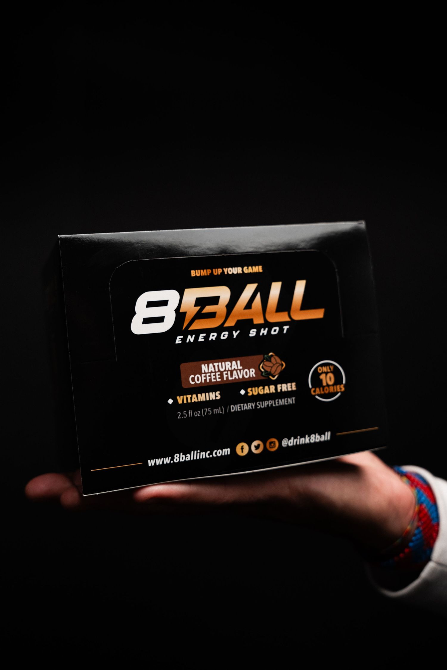 Hand holding a black box of 8Ball energy shot with natural coffee flavors and vitamins against dark background.