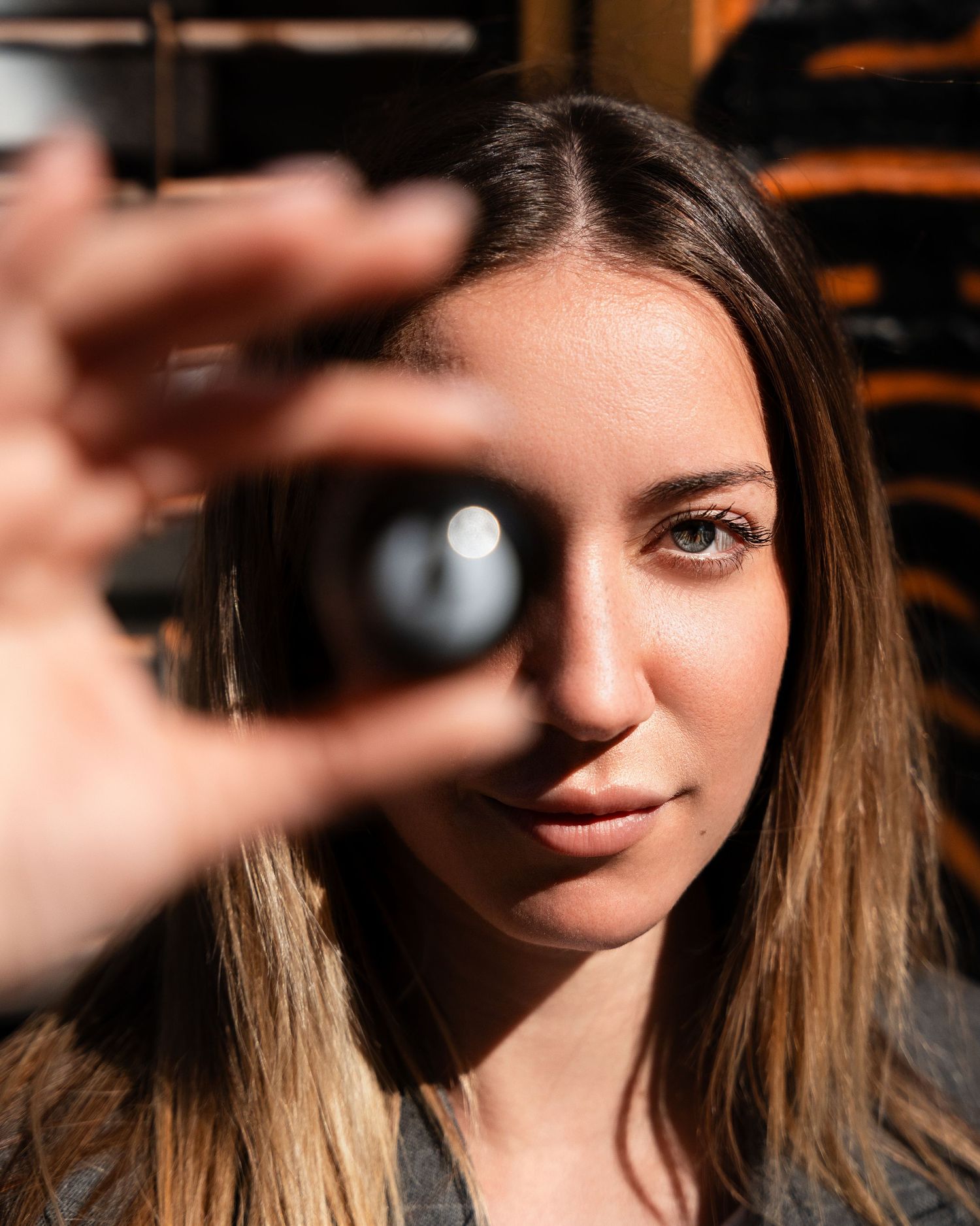 A woman holds up a dark glass orb in front of her face while making direct eye contact with warm natural lighting.