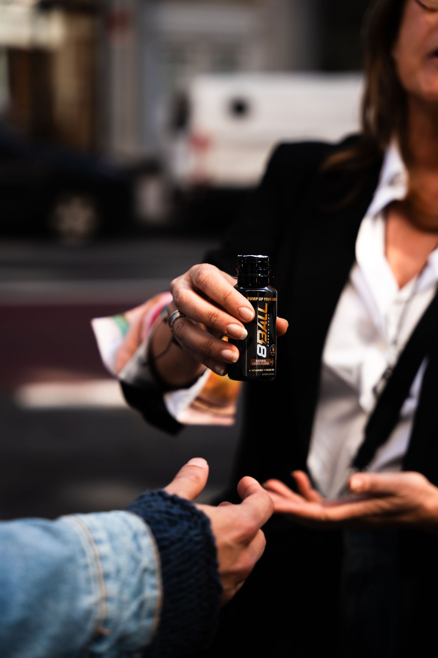 Two hands exchanging a black energy drink can in a street scene with selective focus on the product.