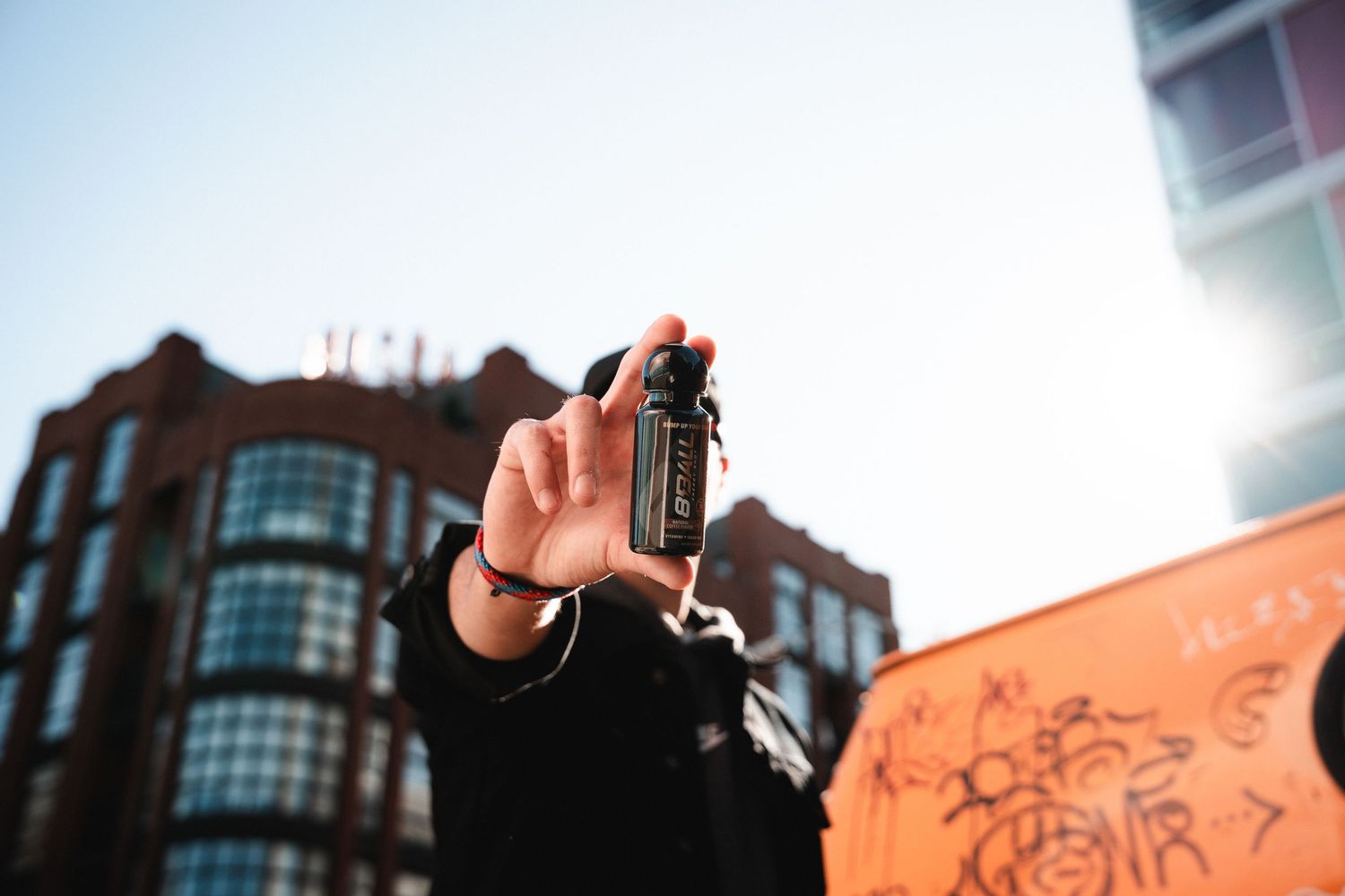 Hand holding a small bottle against urban backdrop with buildings and sky.