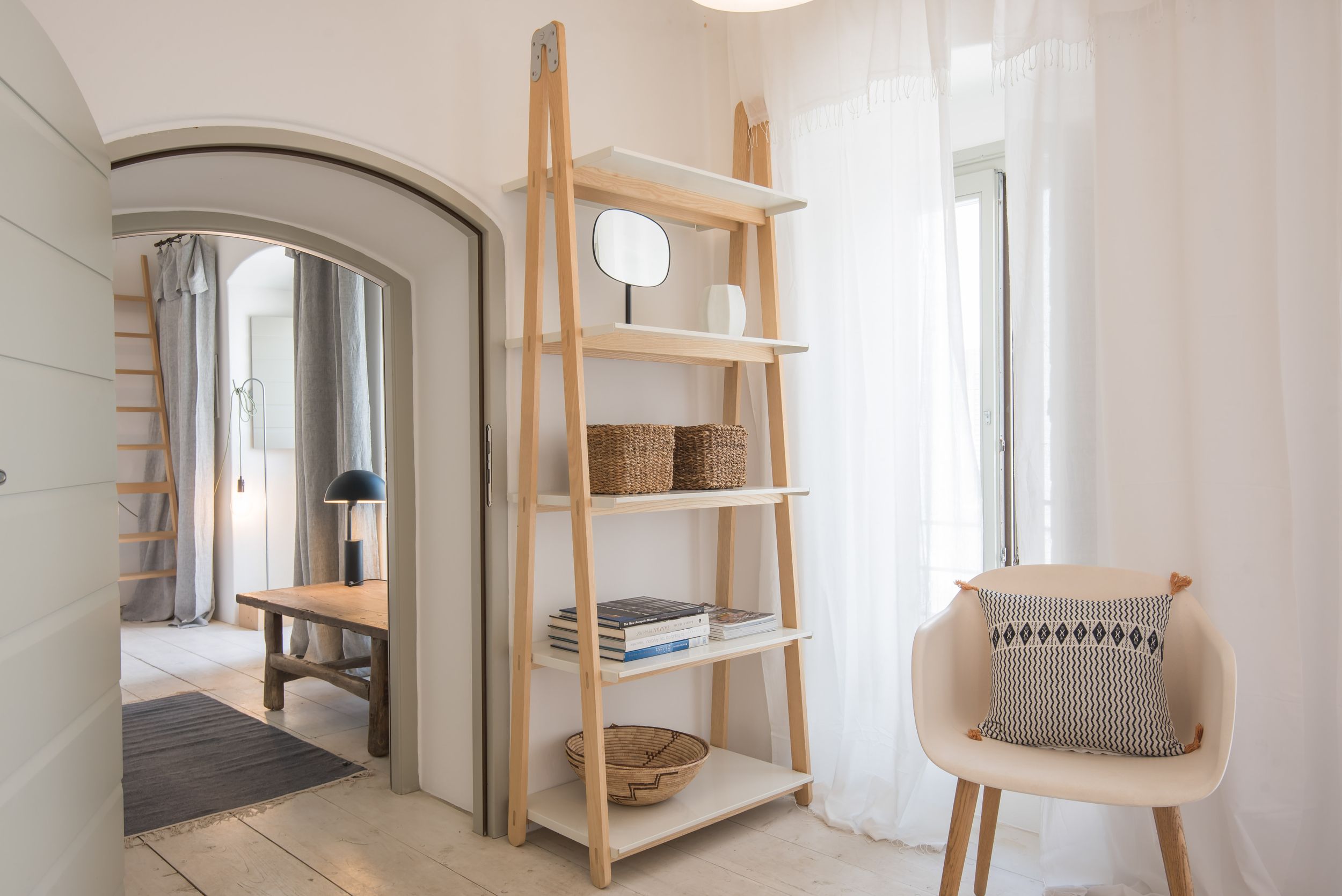 Modern minimalist interior with wooden ladder shelf, arched doorway, and light beige chair against white walls.