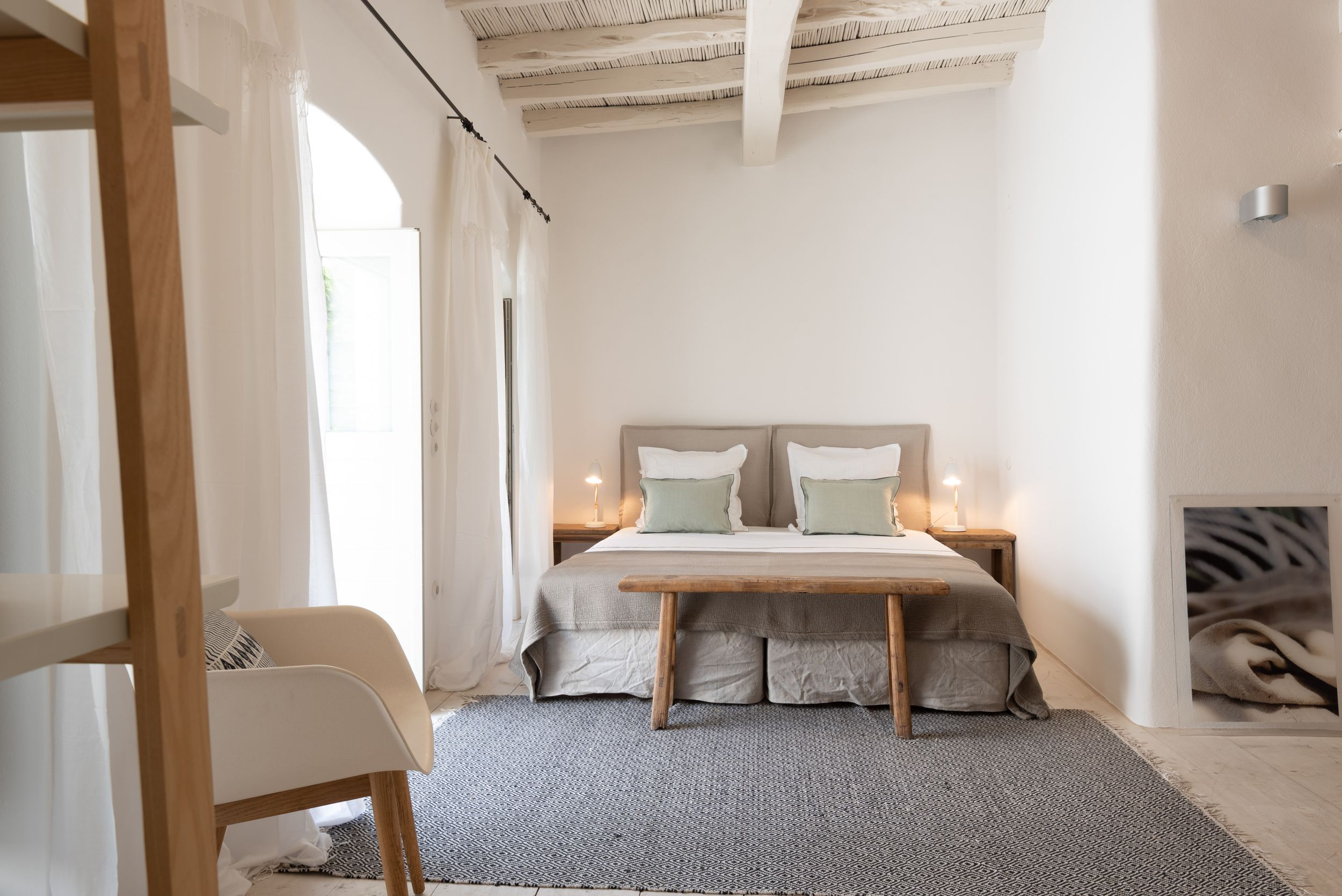 Minimalist bedroom with wooden bed frame, gray bedding, white walls, beamed ceiling, and modern design elements.