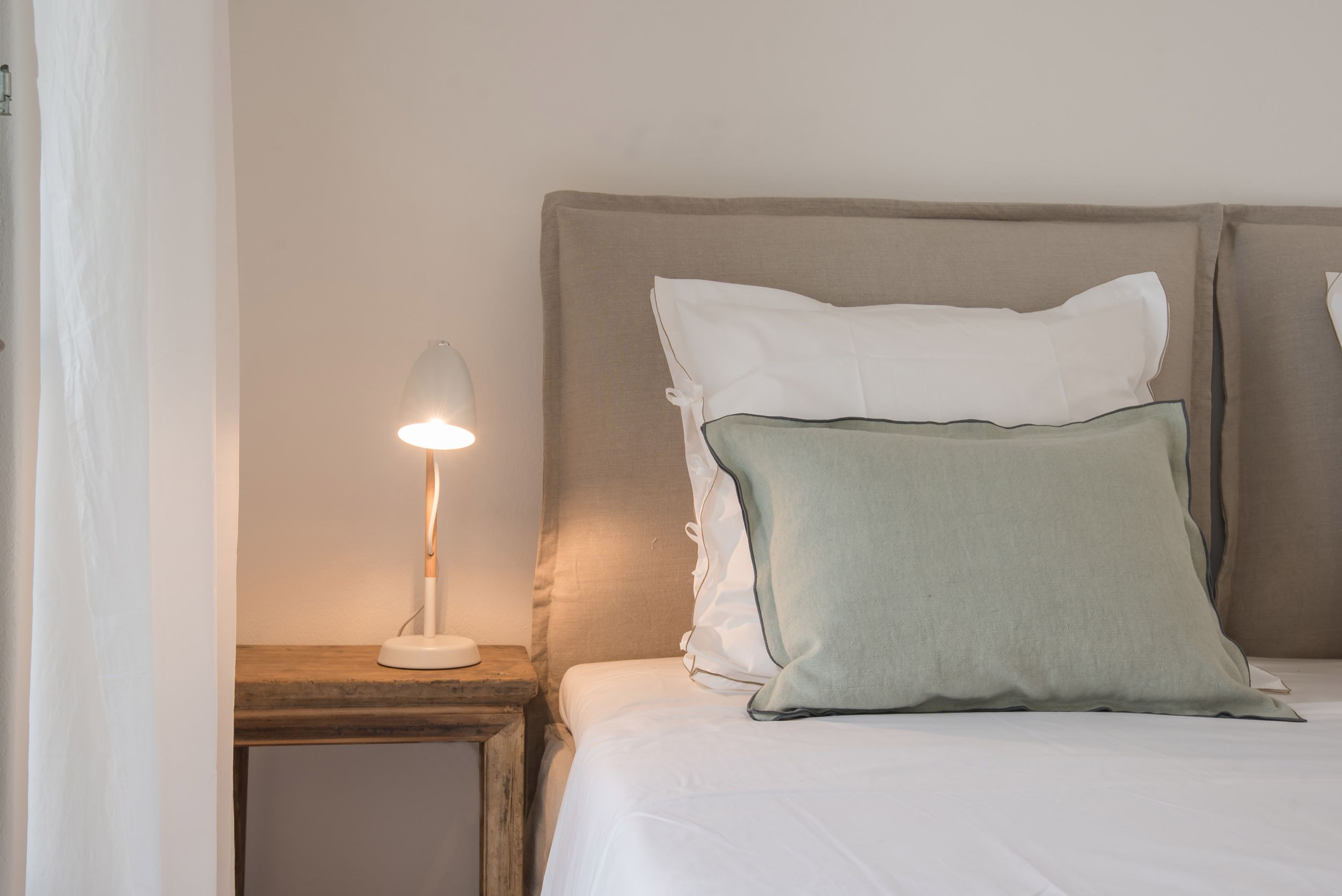 Cozy bedroom corner with white and sage green pillows on gray headboard bed next to wooden nightstand with table lamp.