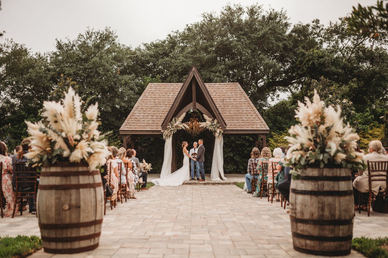 Rustic outdoor wedding ceremony setup with wooden chapel and wine barrels decorated with pampas grass arrangements.