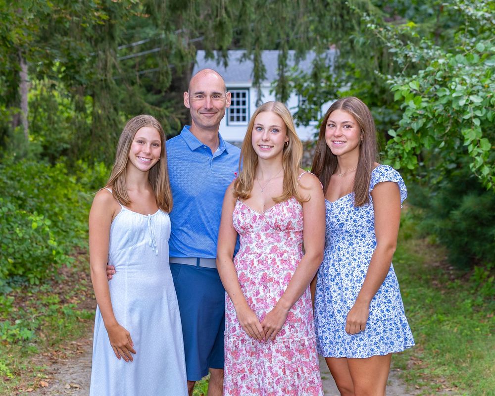 Father & daughters pose outdoors in coordinated blue and pink summer outfits against lush green foliage at high school senior photos.