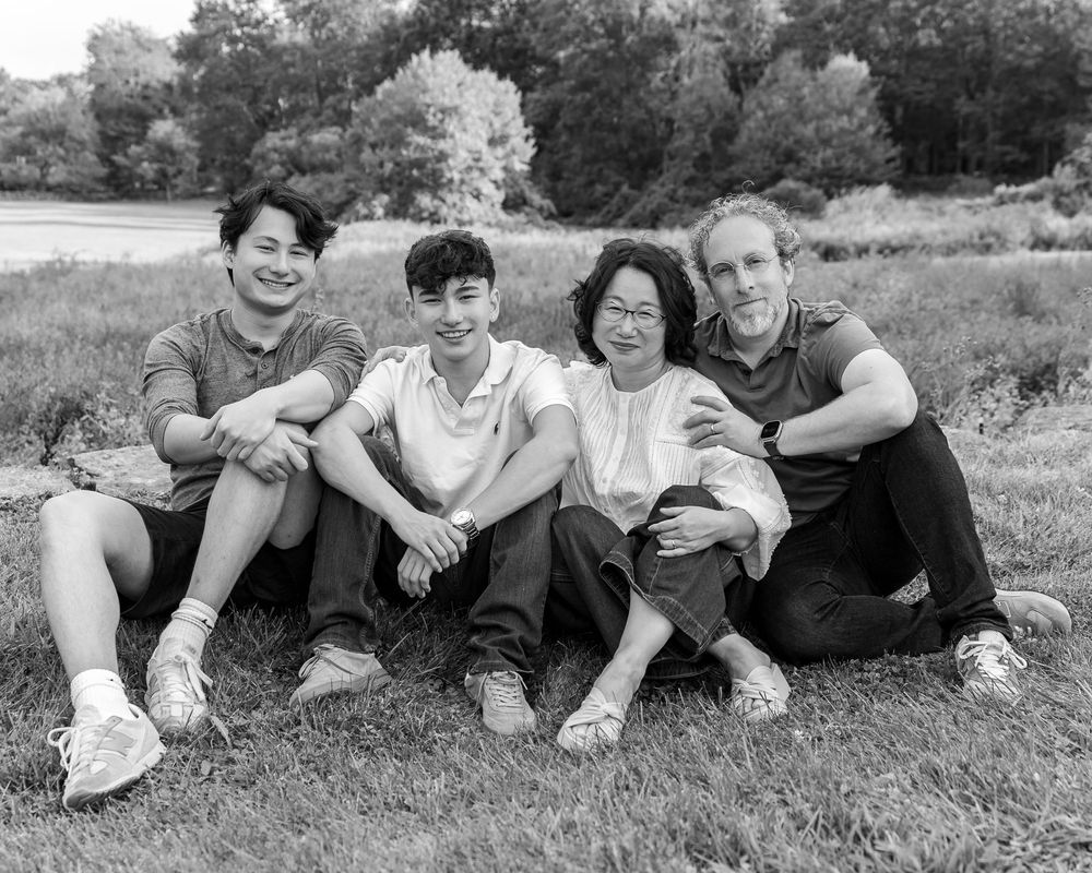 Black & white portrait of a family sitting together on grass with trees in the background during family photography session in Lincoln, MA.