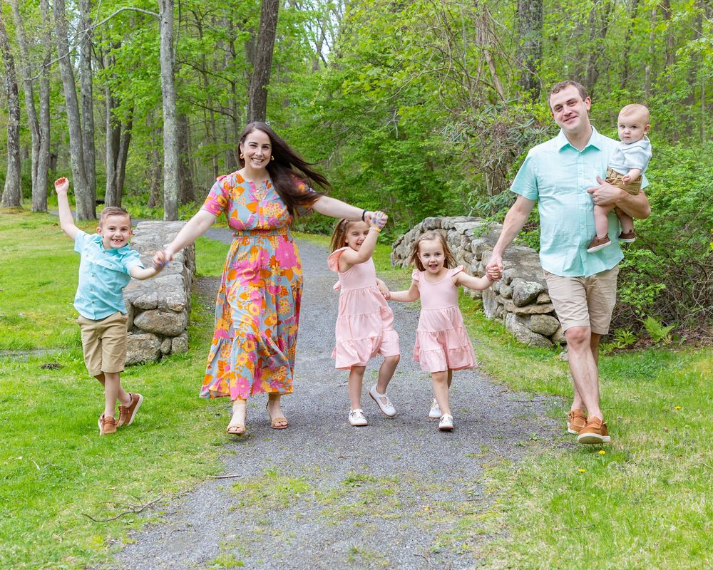 A family walks along a winding forest path surrounded by lush green trees and foliage on a sunny day during a photo session in Sudbury, MA