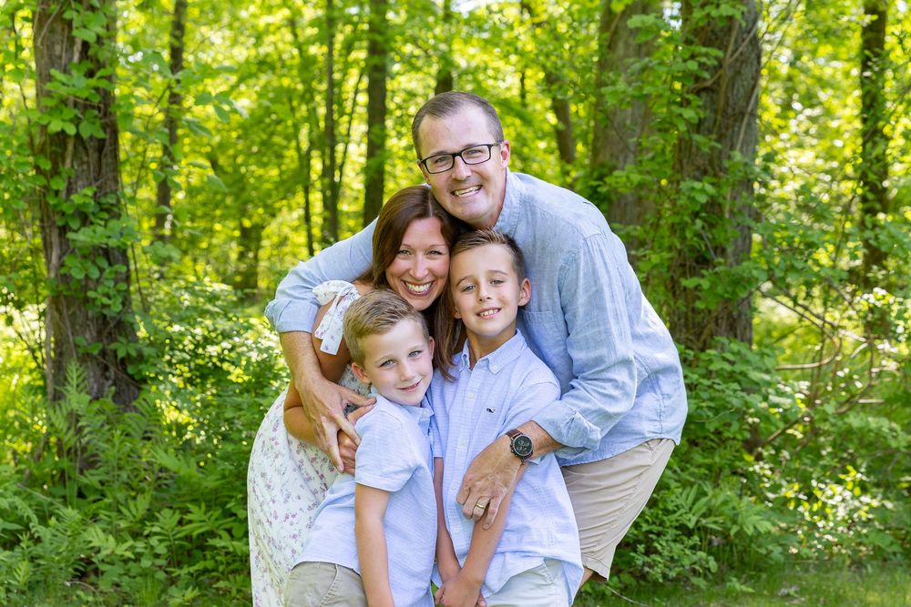 A family of four embraces while posing together in a sunlit forest with lush green foliage in the background in Sudbury, MA.