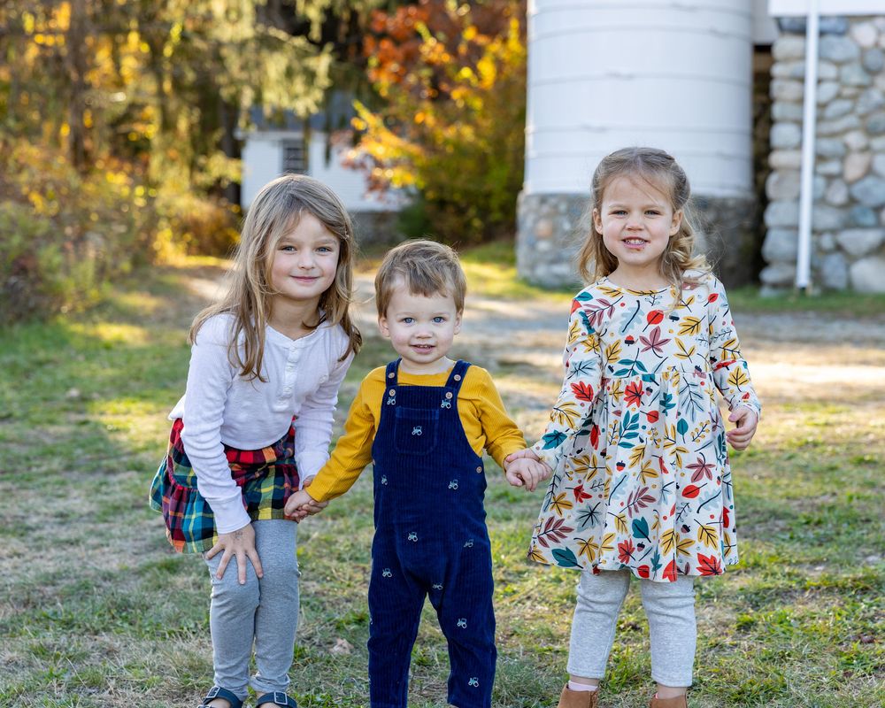 Siblings in casual clothing walk together pose in front of a barn on a sunny autumn day with golden foliage in the background in Sudbury, MA