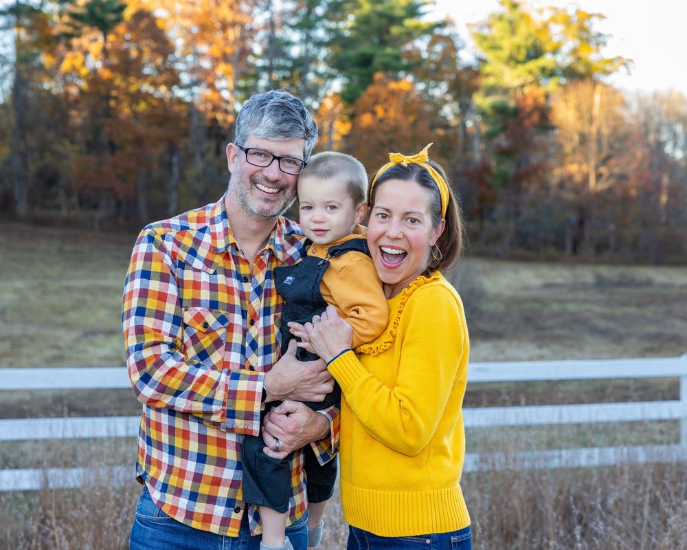 A family shares joyful moments together in a fall setting with white fences and autumn foliage in the background at family photo shoot