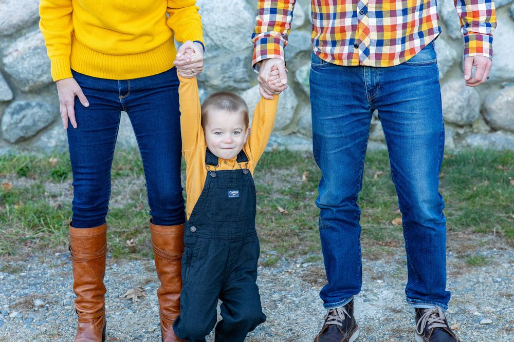 A small child in denim overalls holds hands with two people while standing outdoors against a stone wall during a family photography session