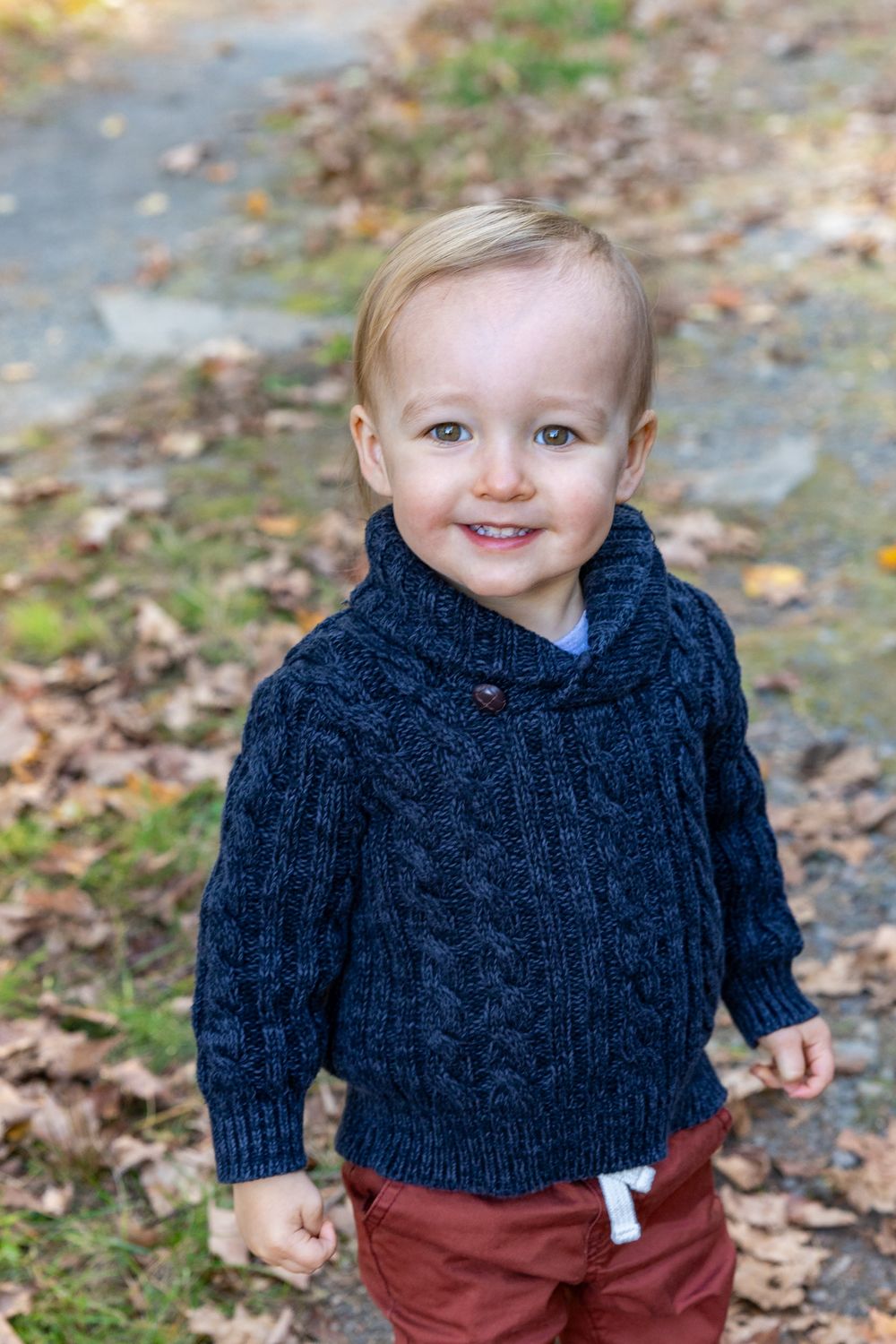 Toddler boy stands smiling among fallen autumn leaves during a photo session in Sudbury, Massachusetts in Metrowest of Boston.