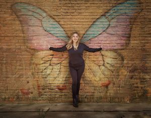 A figure stands against a brick wall with colorful butterfly wings painted behind them creating an interactive mural effect.