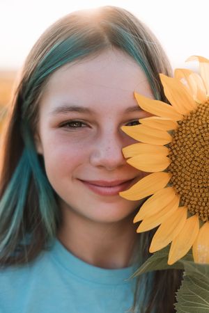 A bright yellow sunflower petal frames a smiling face with teal-colored hair against a sunny outdoor background.