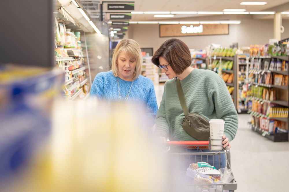 Two shoppers browsing items in a grocery store aisle with shelves of products visible.