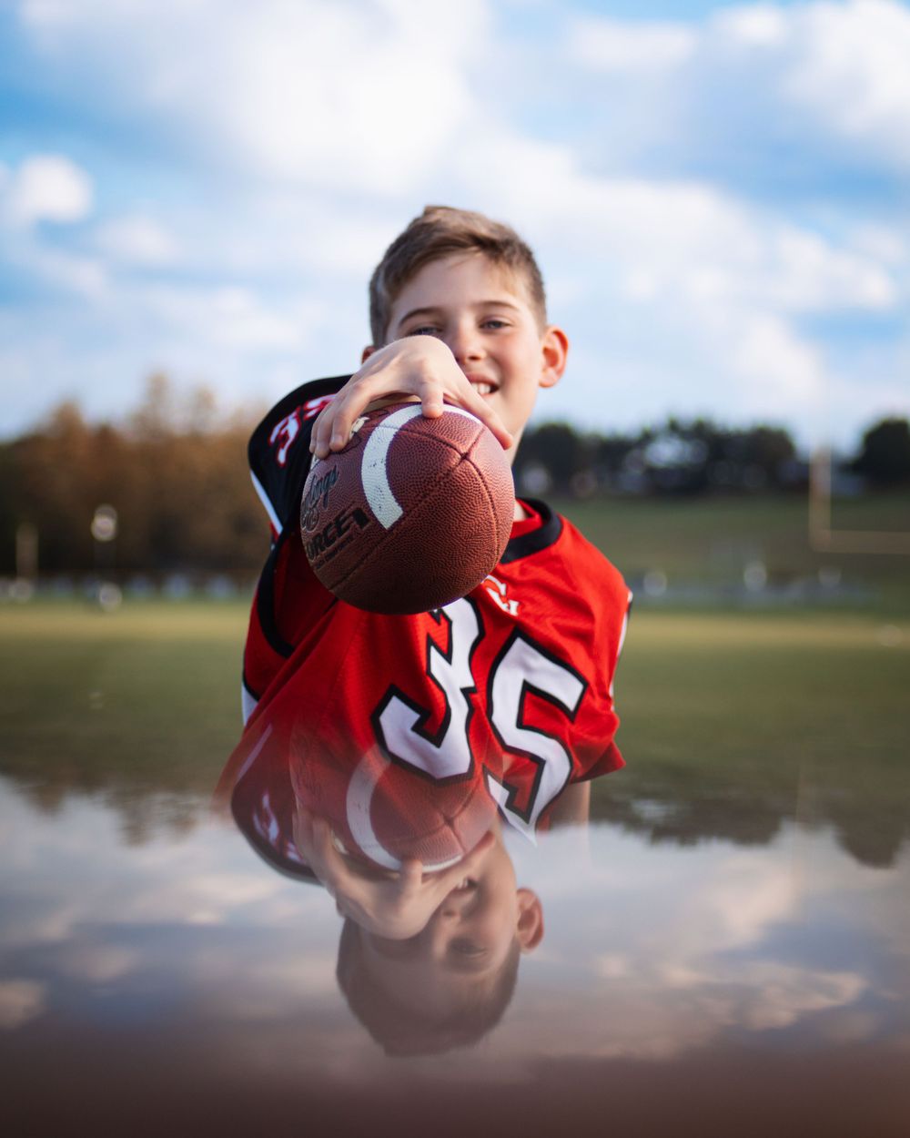 Young athlete in red jersey number 35 holds football with reflection in puddle below.
