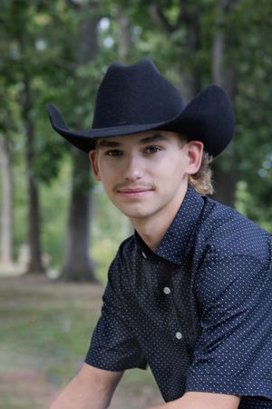 Young person wearing a navy blue cowboy hat and polka dot button-up shirt poses outdoors among trees.