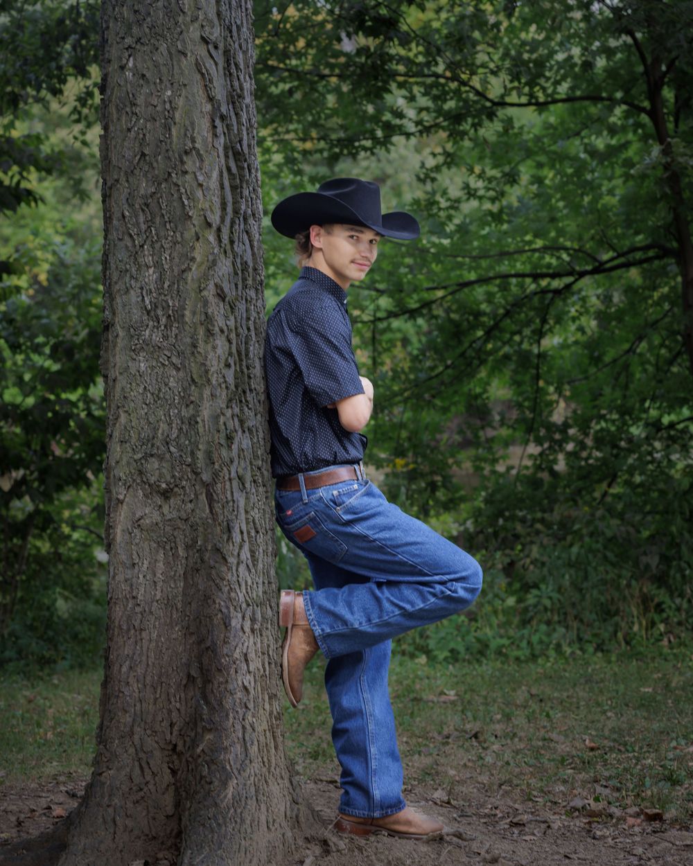 A small child in overalls and a hat climbs up a large tree trunk in a wooded outdoor setting.