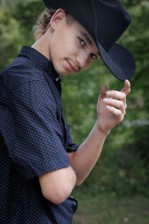 A figure in a navy polka dot shirt and dark baseball cap poses against a blurred green outdoor background.