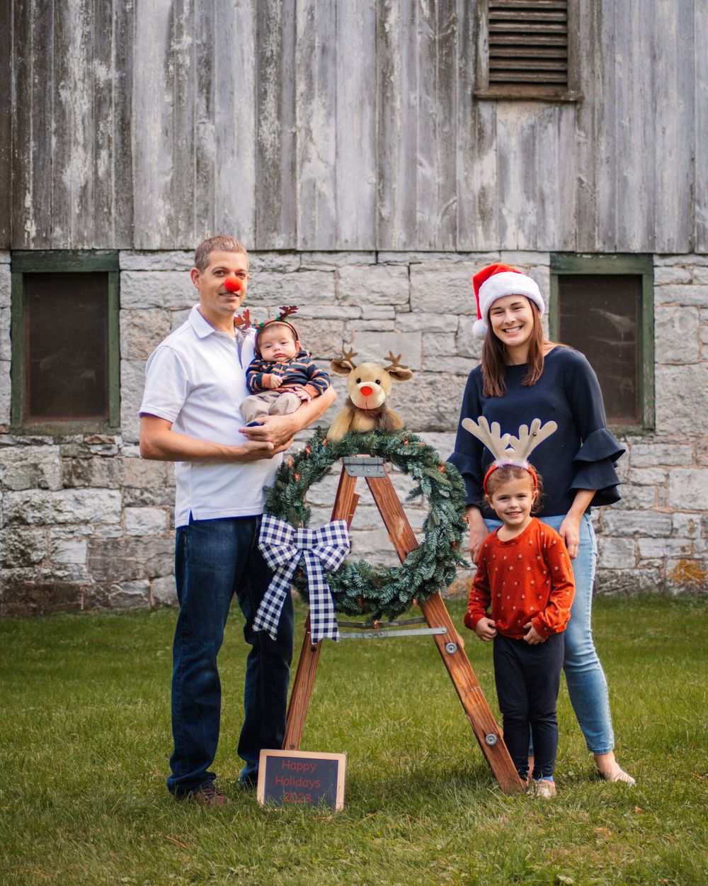 Family poses outdoors with holiday wreath and festive decorations against rustic wall.