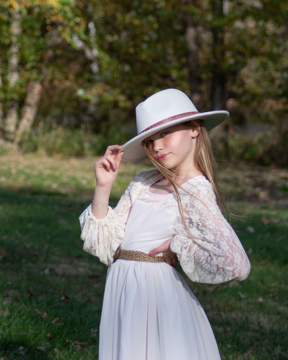 A person in a white prairie dress and wide brim hat poses elegantly in a sunlit field.