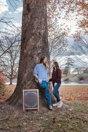 A couple poses near a large tree trunk in autumn with a chalkboard sign during an outdoor photo session.