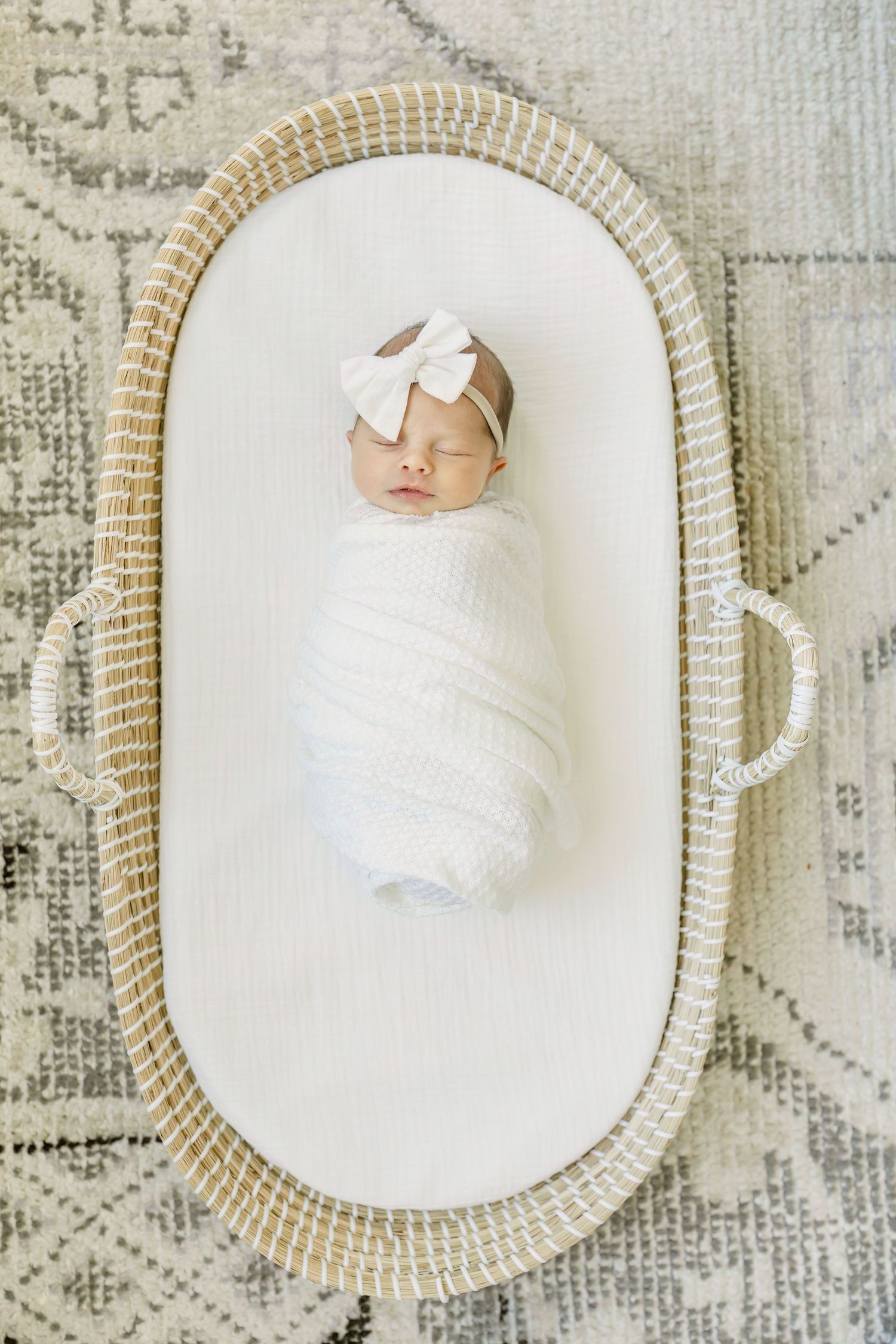 Sleeping newborn wrapped in white with bow headband in woven basket.