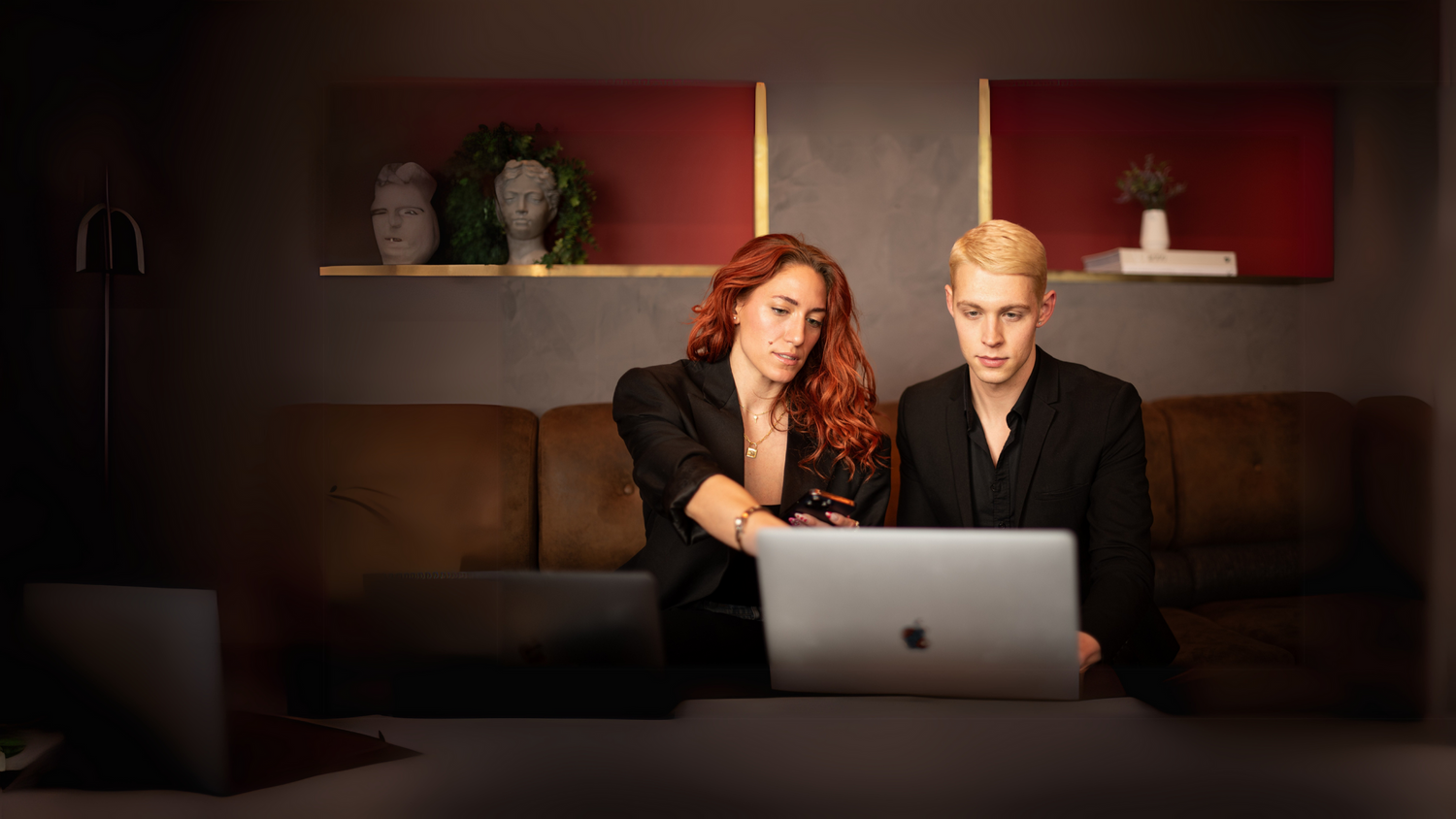 Two people work together while looking at a laptop on couch with red wall and decorative shelves in background.