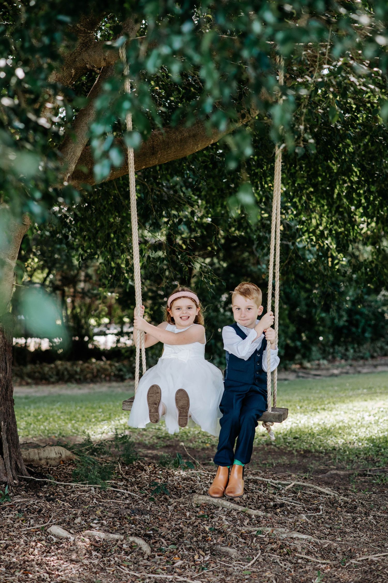 Two children in formal attire playing on tree swings in a garden.