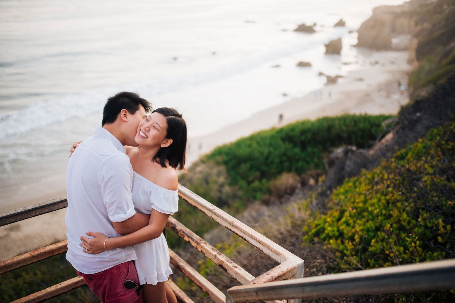 A couple embraces on a coastal overlook with ocean views at sunset.