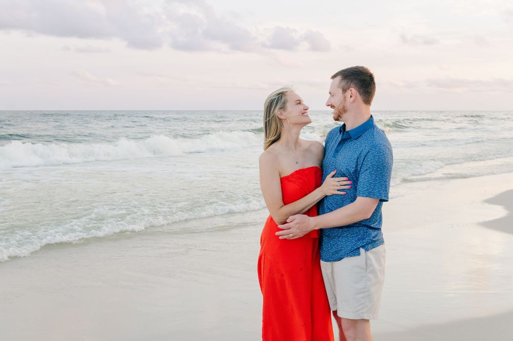Romantic couple embracing on beach at sunset with waves and cloudy sky in background.