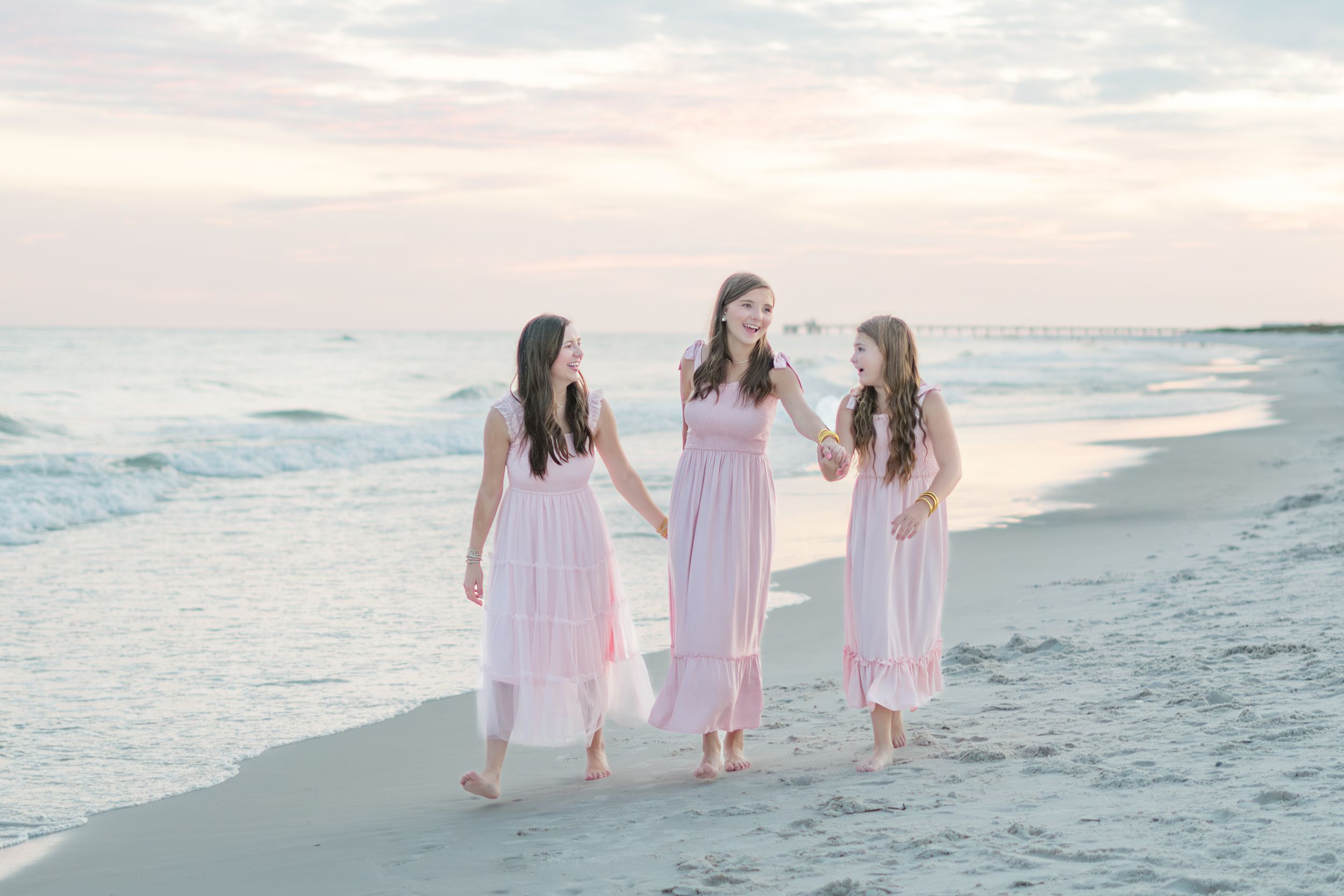 Three people in matching pink dresses walk together along a peaceful beach at sunset.