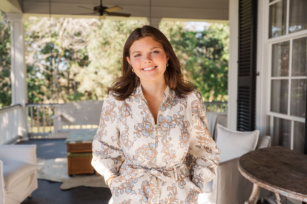 Woman in floral dress smiling while relaxing on front porch of traditional home.