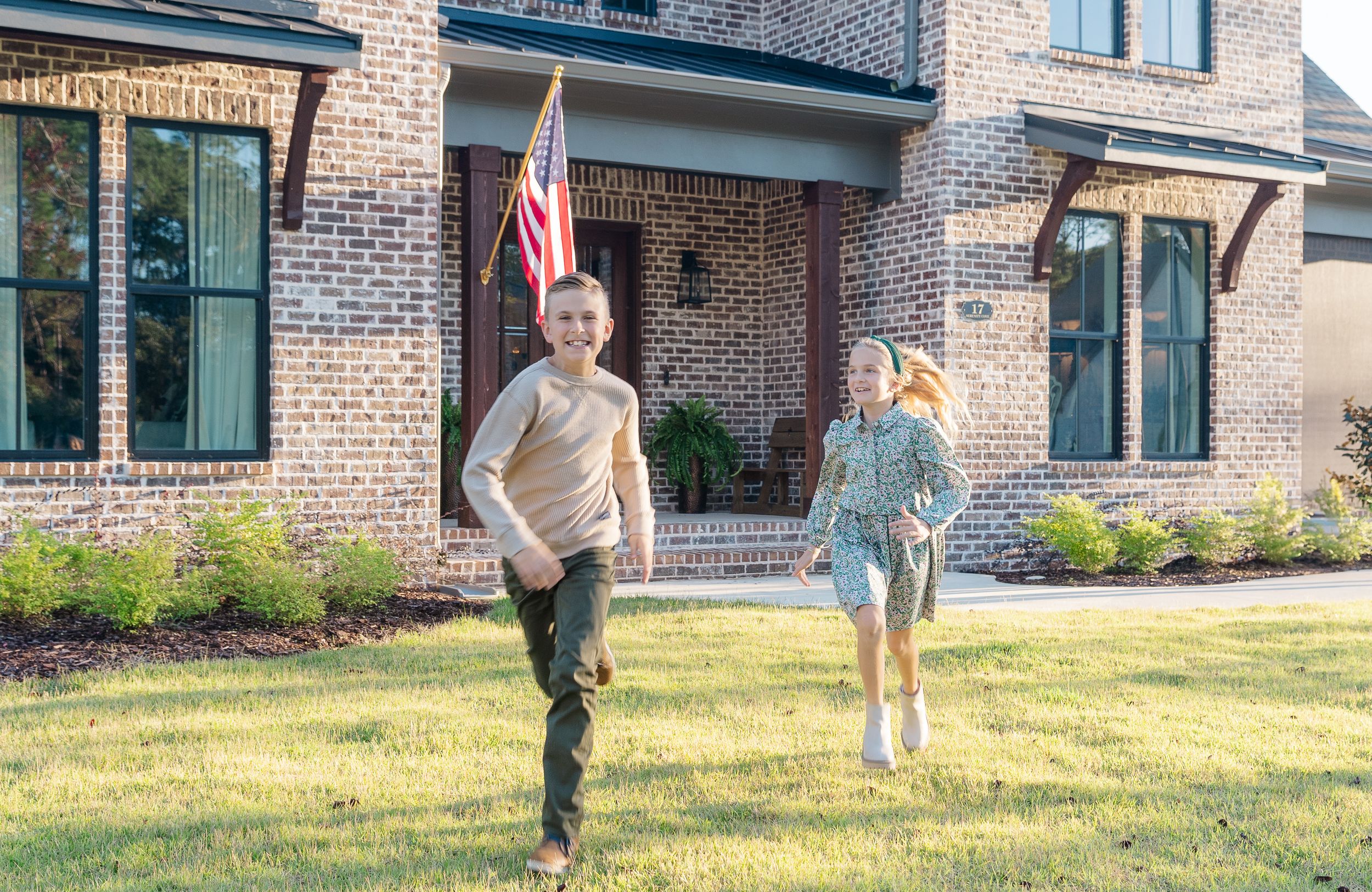 Children playing and running on lawn with American flag in front of brick house.