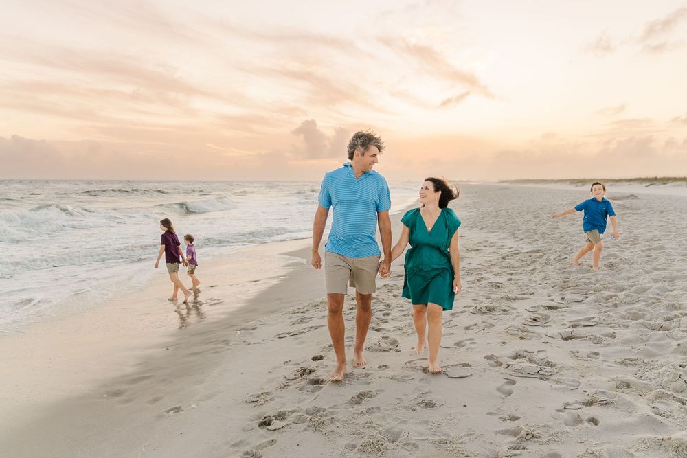 Family walking along beach shoreline at sunset with child running ahead.
