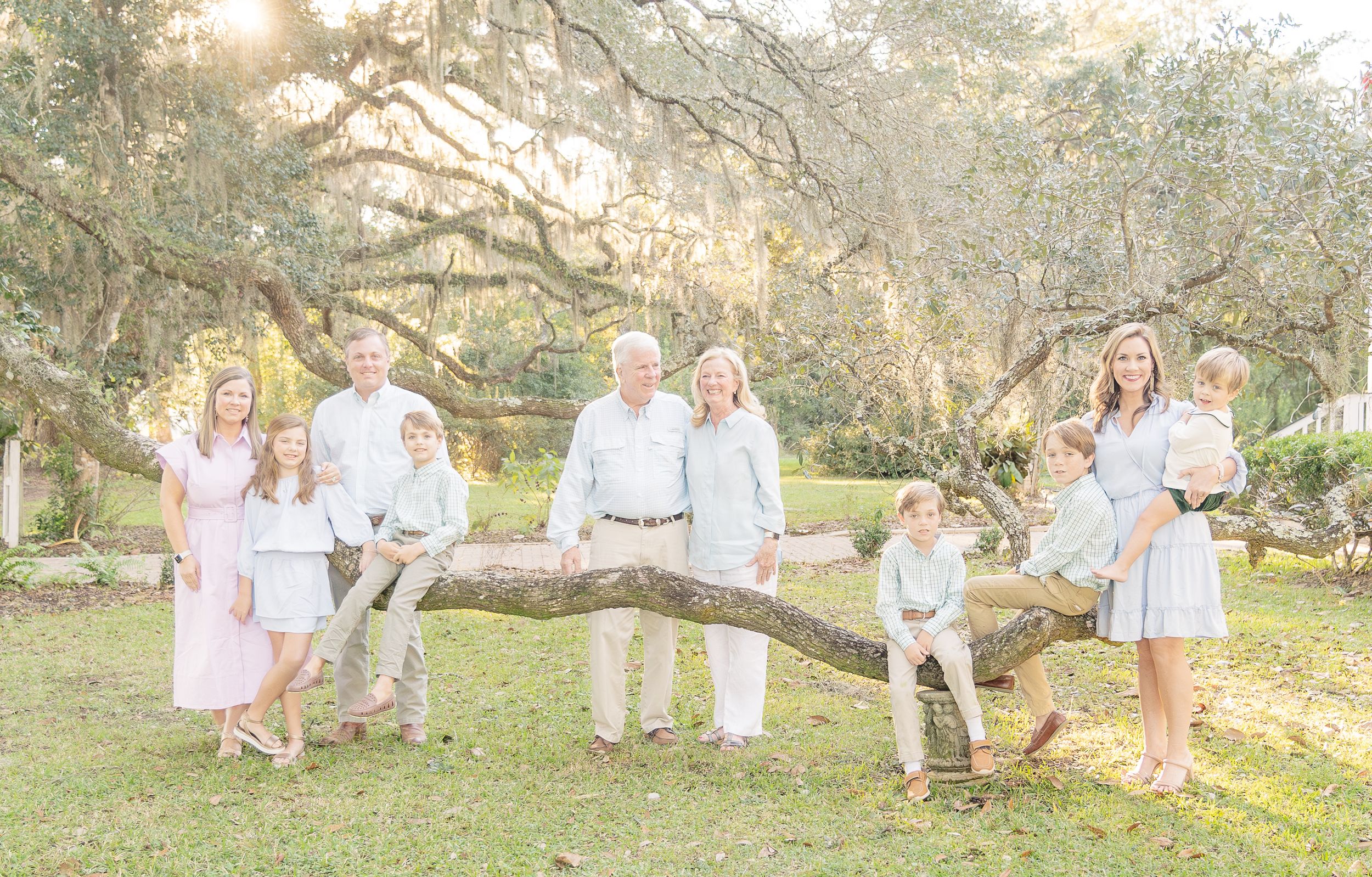 Extended family portrait in white and blue clothing at sunset under oak trees.