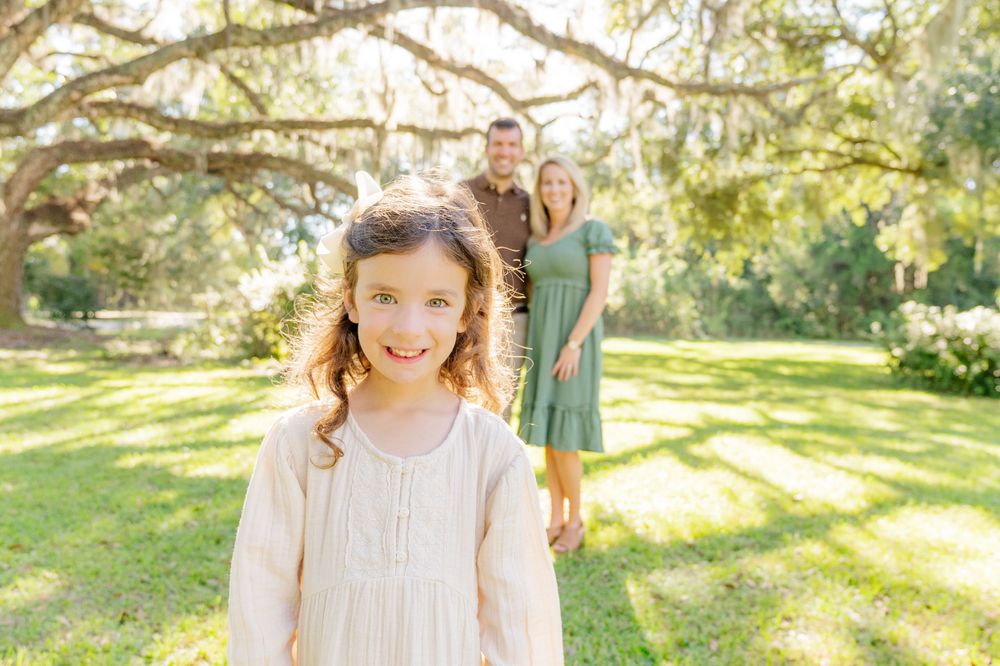 Young girl in cream dress smiling in sunlit garden with parents in background.