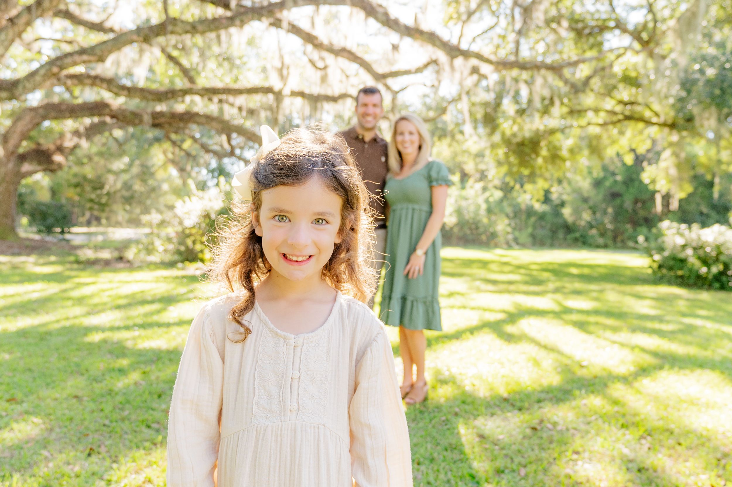 Young girl in cream dress smiling in sunlit garden with parents in background.