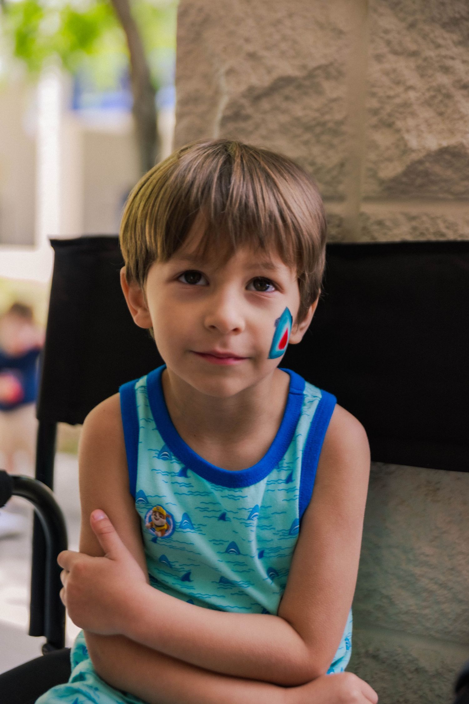 Young child in turquoise tank top with blue face paint sitting in chair outdoors.
