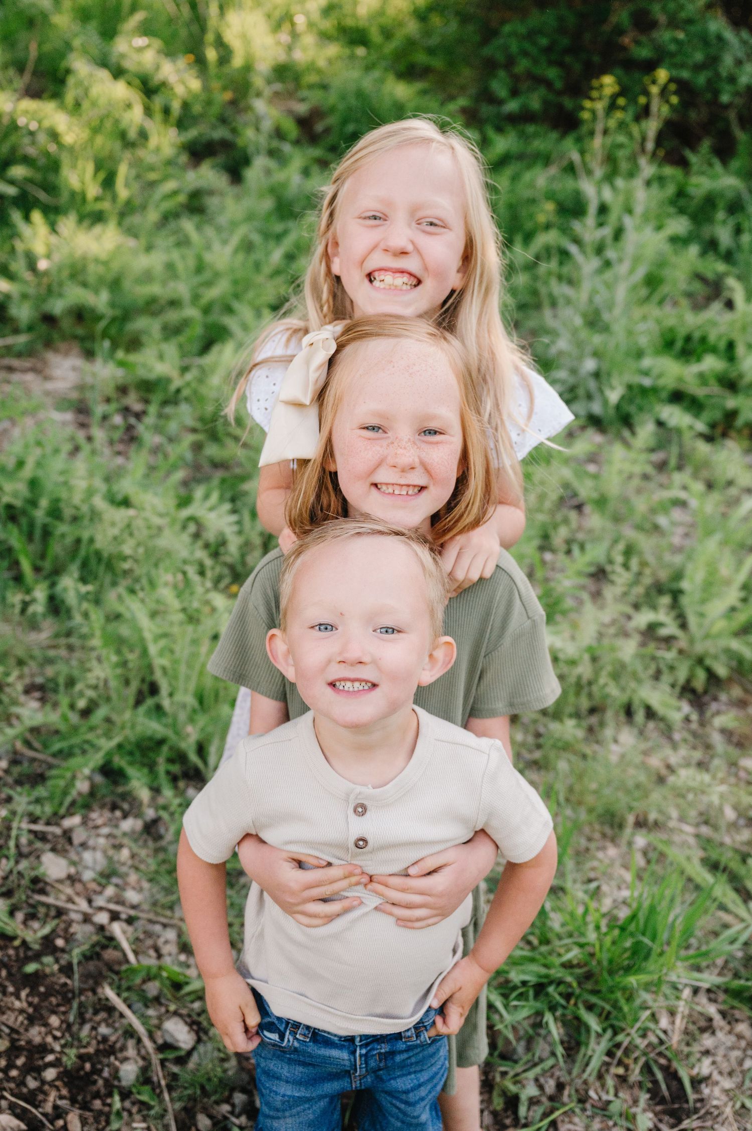 Three young children stand stacked behind each other smiling outdoors with green foliage in the background.