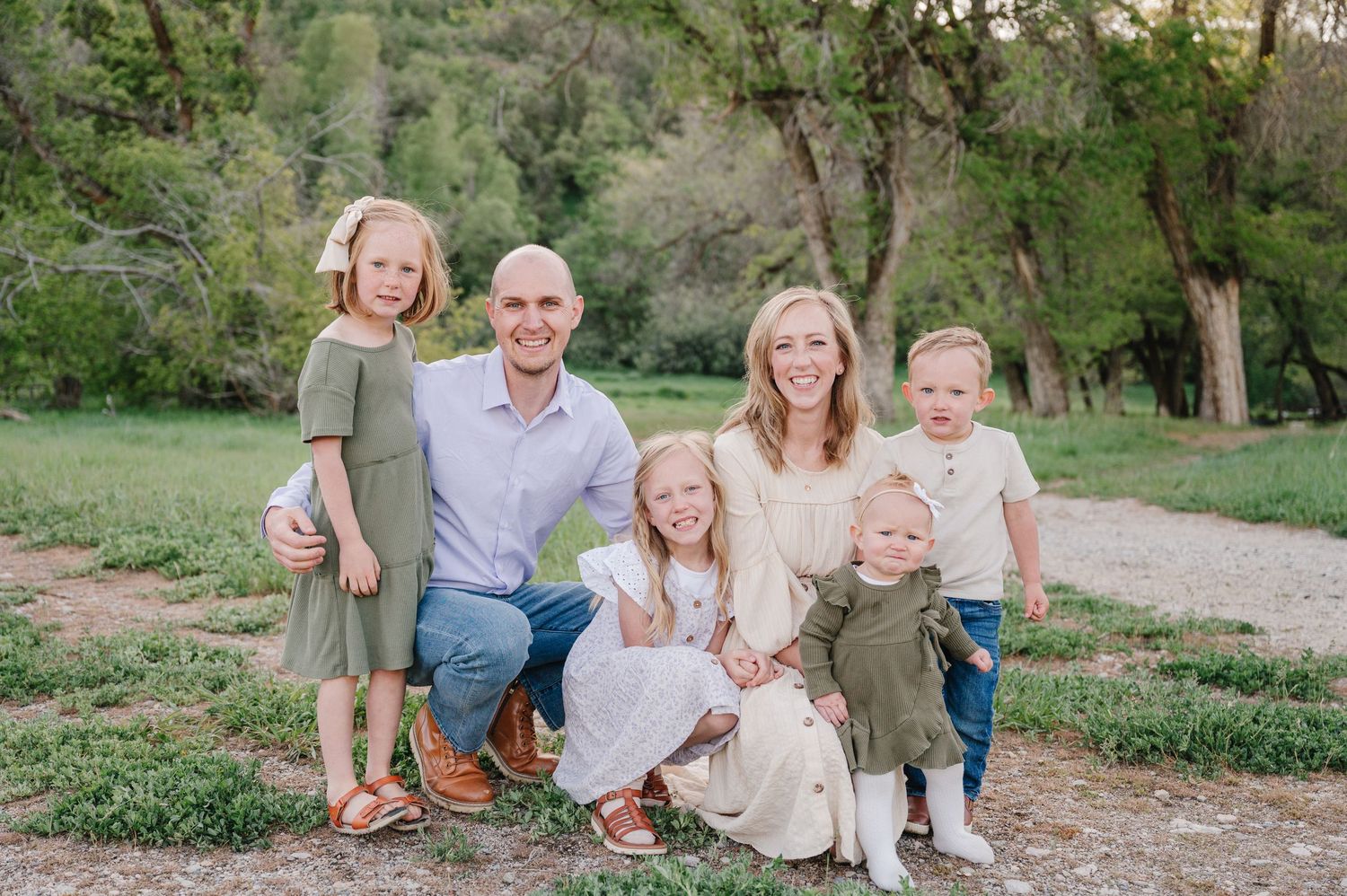 Family group in coordinated casual outfits sitting together outdoors on grassy terrain with trees in background.